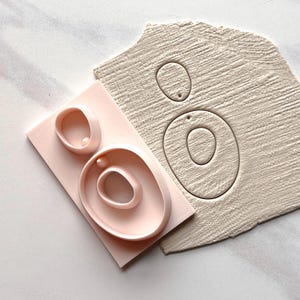 May include: A light pink polymer clay earring cutter set with three different sized circular shapes. Next to the cutters is a piece of textured clay with the same circular shapes pressed into it. The background is a white surface.