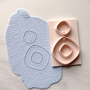 May include: A light blue clay slab with a textured surface and two abstract cut-out shapes. Next to it, a pink rectangular block holds two matching, raised, abstract shapes, each with a small sphere.