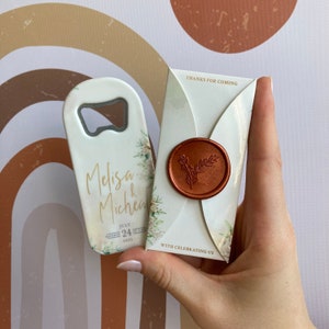 Pode incluir: Um favor de casamento branco com um design floral e o texto "Melisa & Micheal" e "Julho 24, 2023" com um abridor de garrafas anexado. O favor também inclui um envelope branco com um selo de cera e o texto "Obrigado por vir" e "Com nós celebrando".