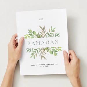 May include: A white book cover with the word "RAMADAN" in gray, surrounded by green and brown leaves. The year "2020" is at the top, and the text "ENJOYING DEEPER CONVERSATION WITH GOD" is at the bottom. The book is held by two hands.