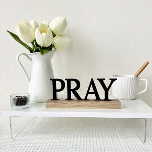 Op de afbeelding: Een witte kan met witte tulpen, een kleine glazen kom, een witte mok met een houten lepel en een zwart "PRAY"-bord op een houten voetstuk zijn op een witte plank geplaatst. De scène speelt zich af tegen een witte achtergrond.