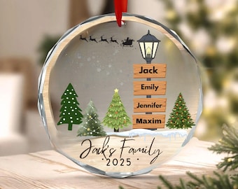 May include: A clear, round Christmas ornament with a red ribbon. The ornament features a winter scene with Christmas trees, a street lamp, and a signpost with names. The text "Jack's Family 2025" is also visible.