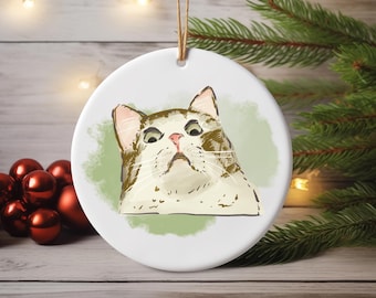 May include: A round, white ceramic ornament featuring a cat illustration. The cat has brown and white fur, pink ears and nose, and a slightly grumpy expression. A green watercolor-style background surrounds the cat. A brown string is attached for hanging.