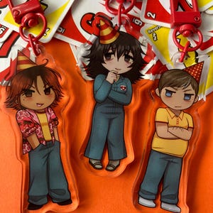 May include: Three cartoon character keychains with party hats. The characters are wearing different outfits, including a blue jumpsuit, a yellow shirt with a floral pattern, and a yellow polo shirt. The keychains are made of clear acrylic and have a red and white Uno card design.