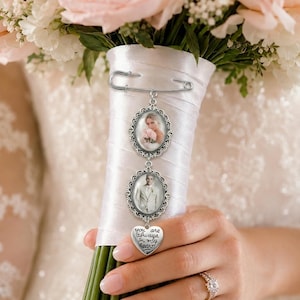 Puede incluir: Un broche de imperdible plateado con dos marcos de fotos ovalados con bordes de filigrana ornamentados y un dije en forma de coraz&oacute;n grabado con el texto "you are always in my heart!" sujeto a un mango de ramo envuelto en sat&eacute;n blanco.
