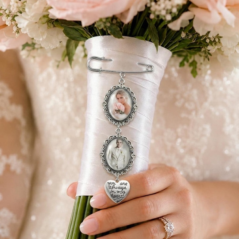 Puede incluir: Un broche de imperdible plateado con dos marcos de fotos ovalados con bordes de filigrana ornamentados y un dije en forma de coraz&oacute;n grabado con el texto "you are always in my heart!" sujeto a un mango de ramo envuelto en sat&eacute;n blanco.