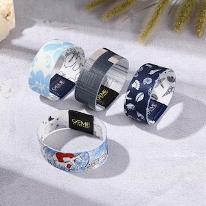 May include: Four fabric bracelets with various patterns. One has a blue floral design, another has a striped pattern, and a third features seashells. The fourth bracelet has a horse and cityscape design. Each bracelet has a black tag with the text "GEME AL JEWELRY".