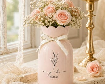 May include: A light pink ceramic vase with a scalloped rim, tied with a cream ribbon, holds a bouquet of pink roses and baby's breath. The vase is decorated with a black botanical illustration and the inscription "j + h 02.15.25".
