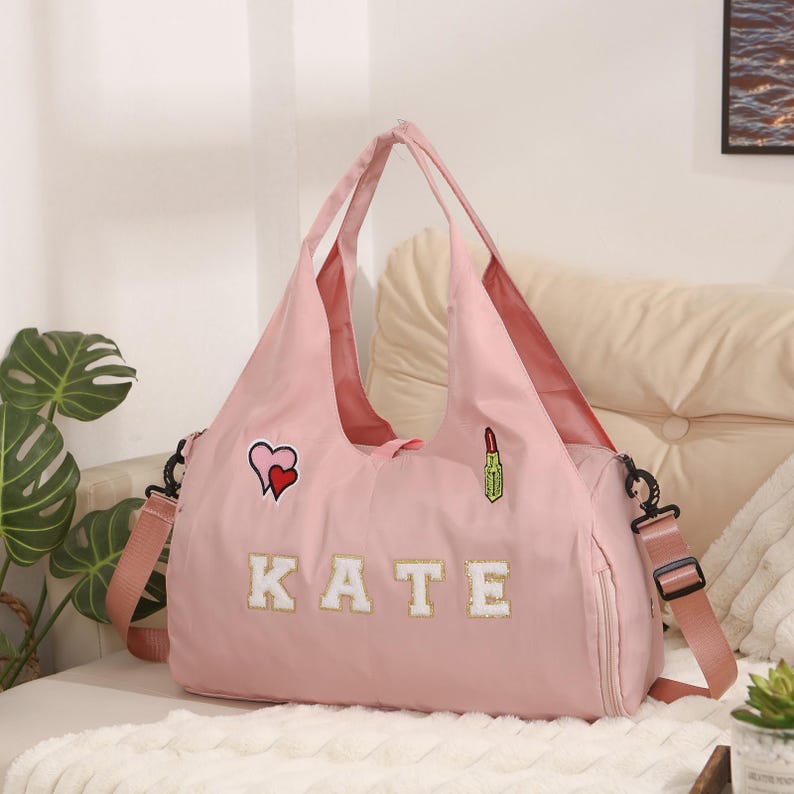 May include: A pink tote bag with a shoulder strap and handles. The bag has the name "KATE" in white letters. It features patches of a heart and a lipstick. The bag is sitting on a white surface.