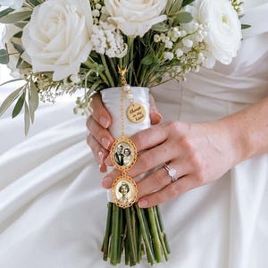 Può includere: Un ciondolo portafoto in metallo dorato fissato a un bouquet di fiori bianchi. Il ciondolo presenta due cornici ovali contenenti ritratti in bianco e nero, un piccolo disco rotondo inciso con il testo Always in my heart e perle bianche.