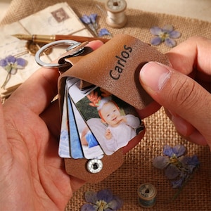 May include: A brown leather keyring featuring a metal ring and a snap button fastening. The leather flap is engraved with the name Carlos. Inside, a small concertina-style photo album displays a picture of a baby wearing a festive headband.