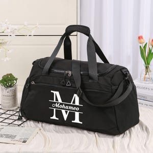 Peut inclure: Sac de sport noir avec un monogramme blanc "M Mohameo IVI". Le sac a une poignée supérieure, une bandoulière et plusieurs compartiments zippés. Une petite plante et des fleurs sont en arrière-plan.