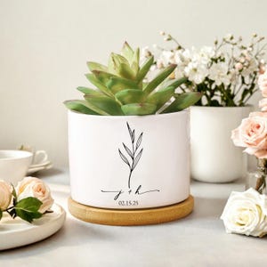 Op de afbeelding: Een witte keramische plantenpot met een vetplant, op een bamboe onderzetter. De pot heeft een zwart botanisch ontwerp en de initialen "j + h" met de datum "02.15.25". Andere witte potten en bloemen staan op de achtergrond.