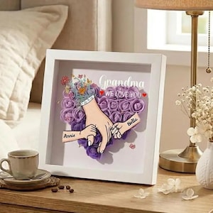 Może przedstawiać: Oprawiona grafika z napisami "Babciu KOCHAMY CIĘ" oraz imionami "Annie" i "Bella". Grafika przedstawia kompozycję w kształcie serca z fioletowych róż, dłonie trzymające się za ręce i bukiet kwiatów.