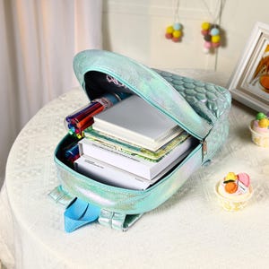 Pu&ograve; includere: Uno zaino iridescente turchese con un motivo a scaglie, aperto per rivelare libri, quaderni e penne colorate. Lo zaino ha spallacci blu ed &egrave; appoggiato su una superficie bianca. La borsa &egrave; progettata per la scuola o i viaggi.