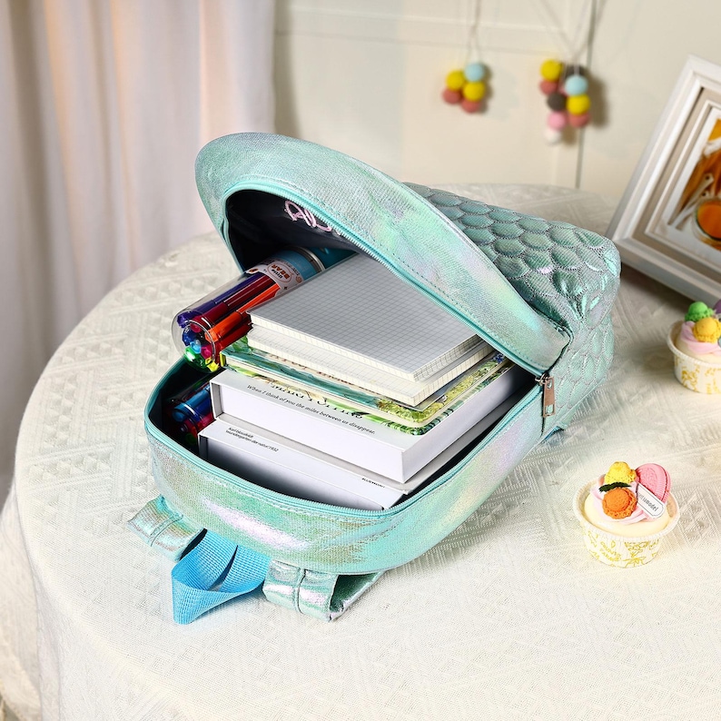 Pu&ograve; includere: Uno zaino iridescente turchese con un motivo a scaglie, aperto per rivelare libri, quaderni e penne colorate. Lo zaino ha spallacci blu ed &egrave; appoggiato su una superficie bianca. La borsa &egrave; progettata per la scuola o i viaggi.