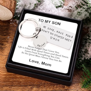 May include: A silver keychain with a keyring and a small tag that says "Skylar". The tag is attached to a larger tag that says "To My Son Be Safe. Have Fun & Don't Do Stupid Shit. ♡ Mom" and "Never Forget That I Love You. Life is Filled With Good Times and Hard Times. Learn From Everything You Can. Be the Great Man I Know You Can Be. Love, Mom".
