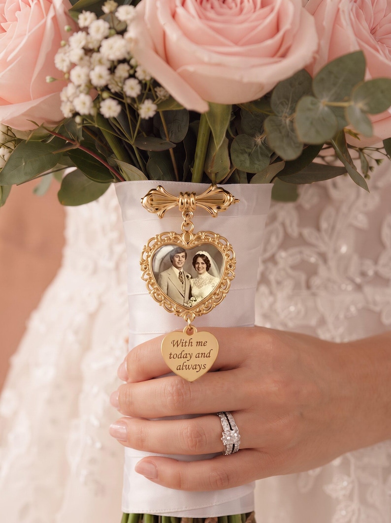 Puede incluir: Un dije de foto de metal dorado sujeto a un ramo envuelto en cinta blanca. El dije presenta un marco ornamentado en forma de coraz&oacute;n con una fotograf&iacute;a antigua de una pareja, y un colgante m&aacute;s peque&ntilde;o en forma de coraz&oacute;n grabado With me today and always.
