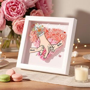 Peut inclure: Une œuvre d'art encadrée en blanc avec un arrangement floral en forme de cœur. L'œuvre présente des illustrations de mains se tenant la main, avec les noms "Annie" et "Bella" sur les mains. Le texte "Grand-mère NOUS T'AIMONS" est également visible.