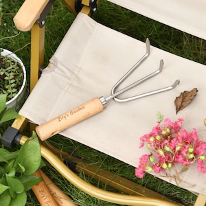 May include: A small garden hand rake with a wooden handle and three metal tines. The handle is engraved with the text "Lily's Garden". The rake is resting on a beige fabric surface, with green grass and pink flowers in the background.
