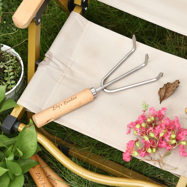 May include: A small garden hand rake with a wooden handle and three metal tines. The handle is engraved with the text "Lily's Garden". The rake is resting on a beige fabric surface, with green grass and pink flowers in the background.
