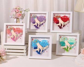 Può includere: Cinque cornici quadrate bianche con facce in vetro, ciascuna contenente una disposizione di rose artificiali colorate a forma di cuore. Le rose sono disposte per formare mani che si tengono per mano, con le parole "Nonna Ti amiamo" in cima a ogni cornice. I nomi "Annie" e "Bella" sono scritti sotto le mani.