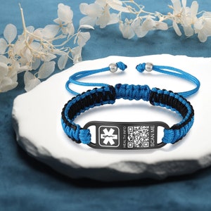 Puede incluir: Una pulsera trenzada azul y negra con una etiqueta de metal negro. La etiqueta tiene un símbolo médico, las palabras "HEALTH INFO" y "SCAN ME", y un código QR.