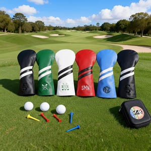 May include: A collection of golf club head covers in black, green, white, red, blue, and black, each with a unique design and initials. Golf balls and tees are arranged on the green grass, along with a small black case with a colorful logo. A golf course is in the background.