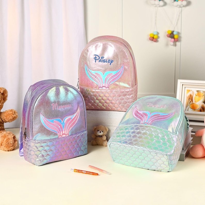 Pu&ograve; includere: Tre zaini iridescenti a tema sirena in rosa, viola e verde acqua. Ogni zaino presenta un design a coda di sirena e un nome in corsivo. Gli zaini hanno un motivo strutturato a forma di scaglie sul fondo.