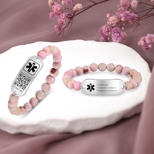Può includere: Due braccialetti di perline rosa con targhette di allerta medica argentate. L'etichetta a sinistra ha un codice QR e il testo "HEALTH INFO SCAN ME". L'etichetta a destra ha un simbolo di stella della vita e il testo "Custom text Telephone number 1 Telephone number 2".