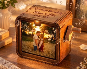 May include: A square wooden music box featuring a hand crank on the side and a photo of a couple in a field on the front. The top is engraved with the text Love keeps the melody Ethan heart Lila 06.12.2019, surrounded by floating musical notes.