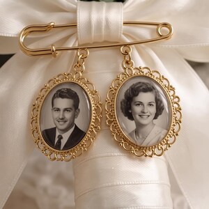 Puede incluir: Un broche de imperdible en metal dorado con dos colgantes ovalados de bordes filigranados. Cada colgante contiene una fotograf&iacute;a de retrato en blanco y negro de una persona. El broche est&aacute; sujeto a un lazo de cinta de sat&eacute;n blanco.