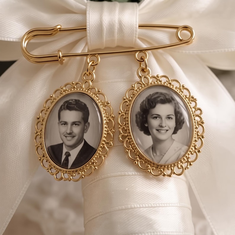 Puede incluir: Un broche de imperdible en metal dorado con dos colgantes ovalados de bordes filigranados. Cada colgante contiene una fotograf&iacute;a de retrato en blanco y negro de una persona. El broche est&aacute; sujeto a un lazo de cinta de sat&eacute;n blanco.