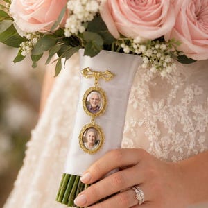 Puede incluir: Un dije conmemorativo de metal dorado con dos marcos ovalados que contienen retratos de un hombre mayor y una mujer mayor, sujeto a una cinta de sat&eacute;n blanco envuelta alrededor de un ramo de rosas de color rosa claro y peque&ntilde;as flores blancas.