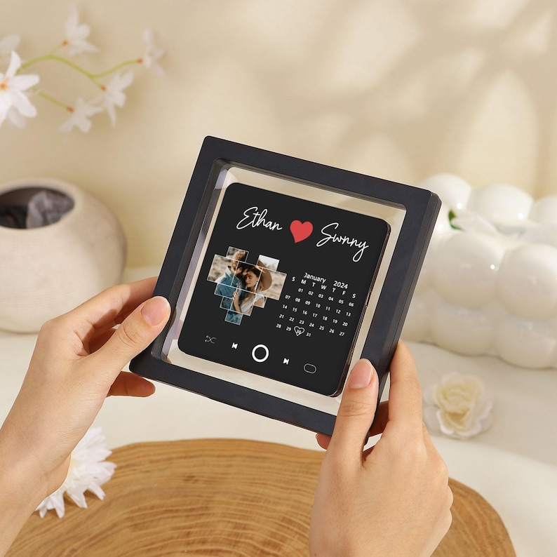 May include: A square black floating frame containing a personalized print with the names Ethan and Sunny separated by a red heart. The print features a photo collage, a January 2024 calendar, and music player interface icons.