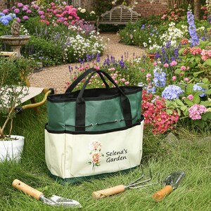 May include: A green and cream-colored garden tote bag with black handles and a drawstring closure. The bag features a floral design and the text "Selena's Garden." Garden tools with wooden handles are in the foreground, set on a grassy lawn.