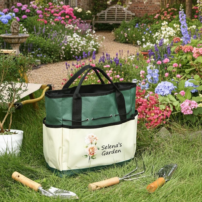 May include: A green and cream-colored garden tote bag with black handles and a drawstring closure. The bag features a floral design and the text "Selena's Garden." Garden tools with wooden handles are in the foreground, set on a grassy lawn.