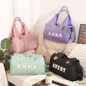 May include: Four personalised duffel bags in pastel shades: pink, lavender, mint green, and black. Each bag has a name in white block letters and decorative patches. The bags have shoulder straps and handles, suitable for travel or gym use.