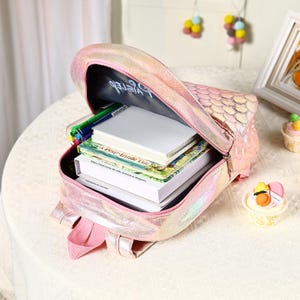 Pu&ograve; includere: Uno zaino rosa iridescente con un motivo a scaglie di sirena, aperto per rivelare libri, quaderni e penne. Lo zaino ha spallacci rosa e una chiusura a cerniera. La fodera interna ha del testo. Una borsa divertente e funzionale.