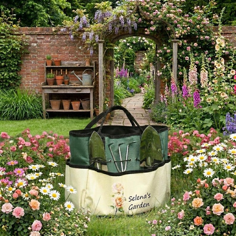 May include: A green and cream-colored garden tote bag with gardening tools. The bag has a black handle and the words "Selena's Garden" printed on the front. The bag is surrounded by flowers and greenery.
