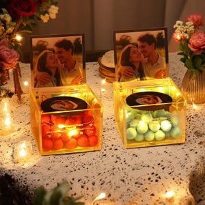 Pode incluir: Duas caixas de exibição de fotos em acrílico iluminadas, uma com esferas vermelhas e outra com esferas verdes. Cada caixa tem uma foto de um casal e um pequeno disco. Luzes de fadas e flores circundam as caixas.