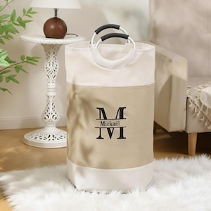 May include: A beige and white laundry hamper with black handles and the name "Mickael" printed in black. The hamper has a circular opening and is sitting on a white fluffy rug.