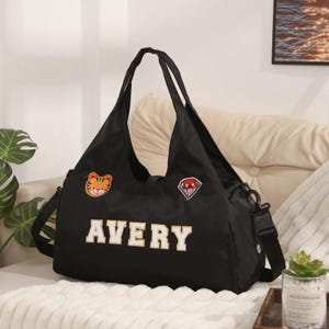 May include: Black tote bag with the name "AVERY" in white block letters. The bag has a shoulder strap and two smaller handles. It features a tiger patch and a diamond patch.