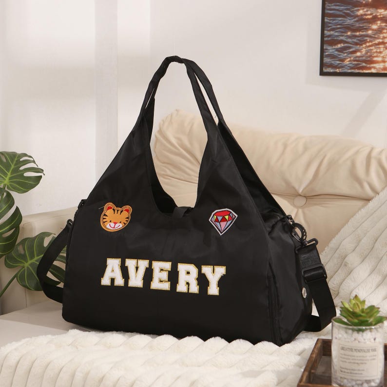 May include: Black tote bag with the name "AVERY" in white block letters. The bag has a shoulder strap and two smaller handles. It features a tiger patch and a diamond patch.
