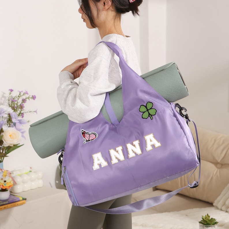 May include: A lilac tote bag with the name "ANNA" in white block letters. The bag features a butterfly and a four-leaf clover patch. A rolled-up green yoga mat is held with the bag. The bag has a shoulder strap and a zip.