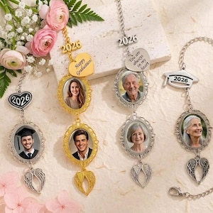 Può includere: Ornamenti fotografici a tema laurea con cornici ovali, ciondoli ad ali d'angelo e l'anno 2026. Alcuni sono dorati, altri argentati. Uno ha un ciondolo a forma di cuore con scritto "Happy Graduation!" e un altro "You are always in my heart."