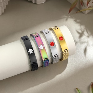 May include: Five medical alert bracelets in black, iridescent, silver, and gold. Each bracelet features a medical symbol in white or red. The bracelets are displayed on a white cylindrical surface.