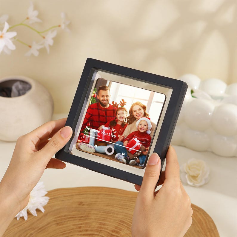 May include: A square black floating frame containing a photograph of a family of four wearing red and white holiday clothing. The photo displays a music player interface with the text Because You Live and the artist name Jesse McCartney.
