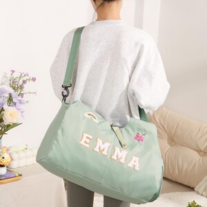 May include: A pale green tote bag featuring the name "EMMA" in white and gold lettering. The bag has a green shoulder strap and a small rainbow and floral patch. The bag is worn over the shoulder.
