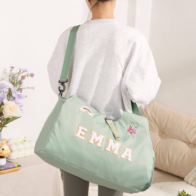 May include: A pale green tote bag featuring the name "EMMA" in white and gold lettering. The bag has a green shoulder strap and a small rainbow and floral patch. The bag is worn over the shoulder.